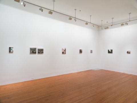 Installation view of nine paintings in the exhibition hung on two walls in a row, evenly spaced apart. The walls are white and the foreground features a warm, brown wooden floor. 