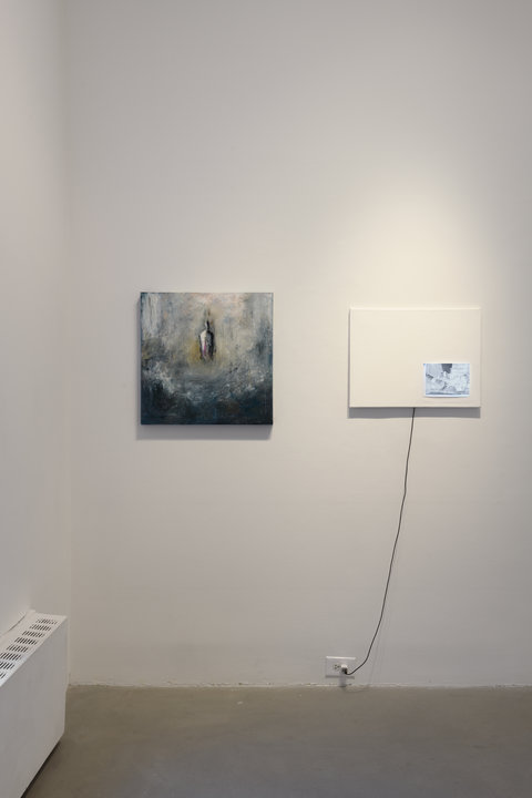 Installation view of two works in the exhibition hanging on a white wall. On the left, an abstract figure is painted into a grey and white abstract background of swirling, and unblended colors. The figure has no face, and no legs, but seems to be emerging from the cloud of painterly smoke. On the right, a white canvas is hung on the wall with a photograph in the bottom right corner. The photograph is negative in color with light shining through to emphasize it. A black cord hangs down from the canvas and plugs into the wall near the ground. 