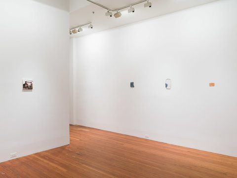 Installation view of several very small, postcard sized works in the exhibition. They are hanging on a white wall, evenly space apart. On the left, a black and white photograph sits besides three works on the right wall. Abstract in material and illegible from the camera view, the works are blue, yellow, and white. 