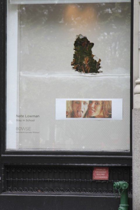 Looking into a large window from the outside. There is a close-up film still of two girls from the movie Thirteen on the bottom right. Above this film still, the wall seems to be broken open to reveal an image of a girl's face surrounded by foliage. On the lower left hand corner, there is text that says "Nate Lowman, Stay in School." 