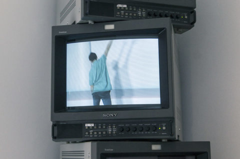 Installation view of a monitor screen in the exhibition featuring a still from a video featuring a person in a bright blue, long sleeve shirt standing with their back to the camera, their right arm pointed up towards the ceiling. Their head is tiled slightly upward.  