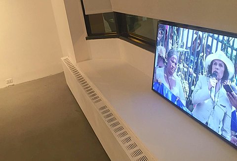 A television monitor showing a figure giving a public speech rest on a window sill in an art gallery.