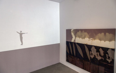 Image of two works from the exhibition on two walls that converge in the corner. On the left wall, a projector projects the image of a white background with a person standing with their arms out wide. On the opposite wall, an abstract painting featuring various strips and sections of dark colors and shapes is hung. 