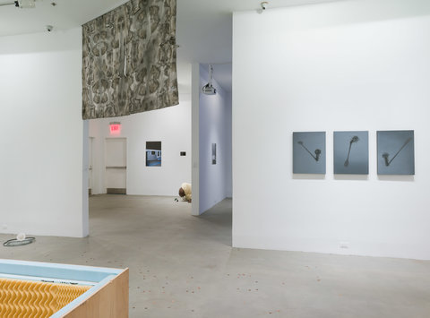 Installation image of several works in the exhibition featuring a hanging tapestry from the ceiling on the left side of the image and three dark grey paintings hanging close together on white wall. 