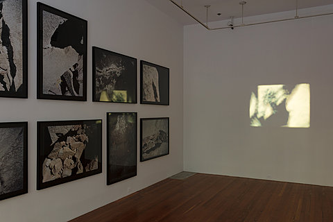 In a dimly lit room there is a projection of an image on the wall directly in view. To the left of the image is an angled view of a wall with black and white photographs. 