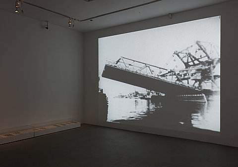 In a dark room, there is a black and white image of a bridge collapsing into water projected on the entire wall.