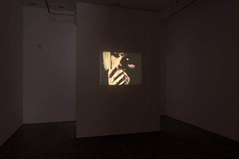 In a dark room there is a projection of a woman's face with her hands covering her mouth in the center of a wall. 