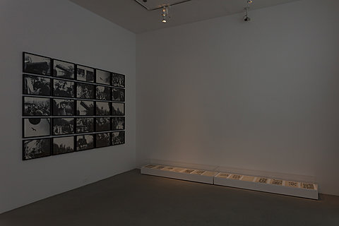 In a dark room on a wall to the left is 25 black and white photographs. The photos take up most of the wall space and are organized in the shape of a rectangle. On the floor in front of the wall on the right is a low glass case which holds multiple warm toned sheets of paper displayed side by side. 
