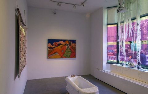 Installation view of the exhibition featuring a bathtub made of paper mache in the center of the gallery. It is white and sits on manufactured claw feet. On the left wall there is a hanging fabric with a projection. On the middle wall, a colorful painting hangs, on the right, pink light streams in through pink film covered windows. 