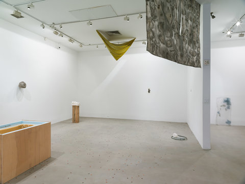 Installation image of several works in the exhibition featuring a hanging tapestry from the ceiling on the left side of the image and three dark grey paintings hanging close together on white wall. 