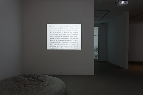 An image of black and white text is projected on a wall directly in the center of a dark room. On the floor in the lower left side is a round and textured grey sculpture