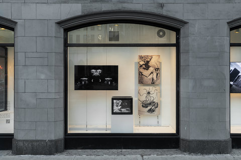 Installation view of the exhibition featuring two monitors on the left, staggered so that the larger monitor is high up towards the left, while the small monitor is low, towards the right. On the right side of the image, two drawings hang vertically on top of one another. They are created in black and white and feature abstract lines and shapes. 