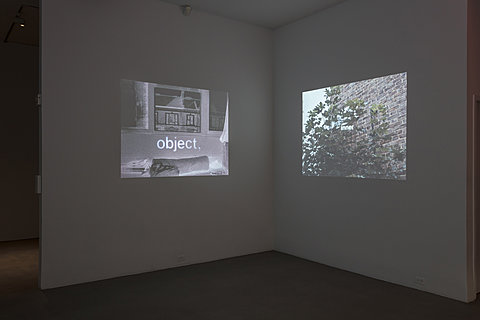 In a dark room, two image projections appear on adjacent walls facing each other. The image on wall to the left is of a window of a building, on bricks under the window is the word "object." written below in white large font. On the right side wall is a muted colored photograph of the side of a building's brick wall. There is a tree with leaves on the left side of the building that takes up nearly the entire picture frame.