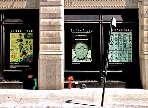 Image of three large windows from the outside with a sidewalk on the foreground. On the first window, there is a yellow poster with various figures. On the second window there is child with a green face and large 'x' on their forehead. On the child's hair is the text 'Have you seen me?" Below there is illegible text. On the bottom of the poster, there is black graffiti that says 'gunk #4.' On the third window there is a poster with small sketches arranged in ten rows of five.  
