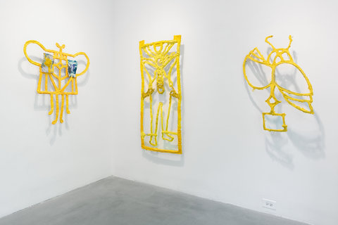 Installation image of three sculptures in the exhibition, mounted to two white walls converging at a corner. On the left wall, a yellow paper mache abstract sculpture is mounted to the wall. In the middle, a yellow paper mache rectangle, with abstract paper mache in side the borders. On the right, another abstract paper mache shape is mounted, with the light reflecting similar, abstract shadows onto the white wall behind it. 