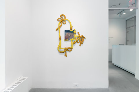 Image of a sculpture mounted on the wall in the exhibition that features a painting with yellow paper mache and cardboard rope protruding from all edges. 