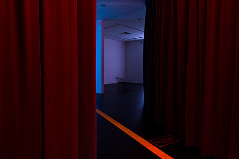 A yellow path on the black floor passes through two parted red curtains to a dark room lit with blue light.