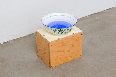Close-up image of a wooden block, placed on the cement floor of the gallery. Atop the block sits a blue, china dish with blue liquid sitting inside. 