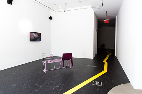 A pink bench faces the corner of the room in which an image of an arm with a wristwatch hangs on the wall. A yellow path on the black floor leads out of the room down a hallway.