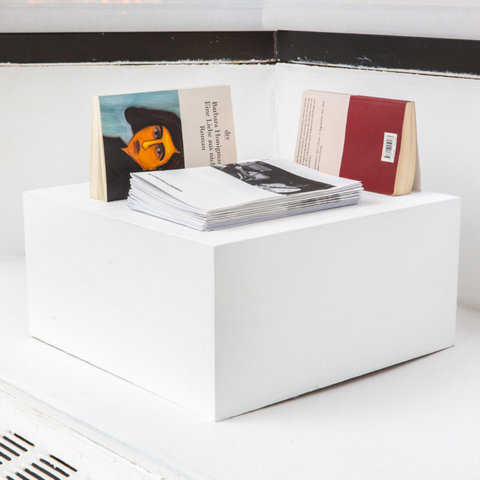Installation image of the exhibition featuring part of the installation set up on the window sill. A small, white cube pedestal sits. Three books sit on top of the pedestal. 