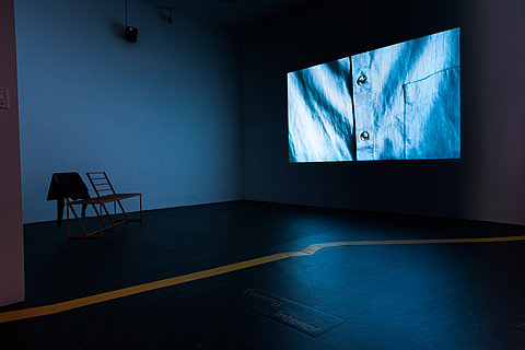 A projection of close up of a button-down shirt is on the wall. There is a bench with a sweatshirt.