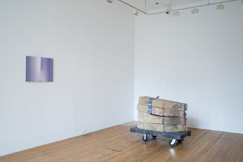 Installation view of the exhibition featuring a purple print hanging on a white wall on the left side wall within the image. On the floor of the gallery, a platform on wheels holds several slabs of what seems to be wood or stone, stacked on top of one another. 