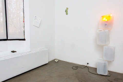 Installation view of the exhibition featuring a sculpture propped against the right-side wall. The sculpture includes several plastic jugs, staggered on top of one another. The one on top, the fourth jug, has a light inside that glows yellow. On the left of the sculpture, a green and white, tie-dye sock hangs on the wall. A small cup sits on the floor in the corner, and a white piece of paper with typed writing, illegible from camera view, is tacked onto the left side wall. 