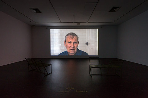 A projection of a figure directly addressing the camera is projected on the white wall, in front of which are two benches.