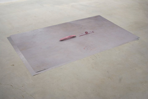 Installation view of a work on the floor of the gallery space. The work seems to be a photograph featuring a pale pinkish, purple background, almost grey with two pinkish-red objects laying in the center. The objects are small rectangular objects, illegible from the angle of the camera and the artwork in general. 