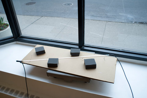Installation image of a work sitting on the window sill of the exhibition. Several cube-shaped pieces of black foam sit atop a rectangle of cardboard. 