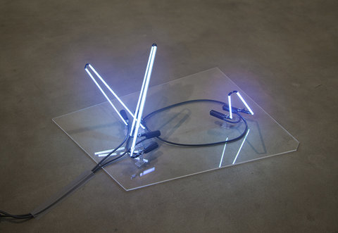 Close-up image of a neon light installation on the floor, in the center of the gallery.In the center, on the floor of the gallery is a neon light installation sitting on a clear rectangle. The neon is blue and two longer beams sit across from two much shorter beams of neon. 