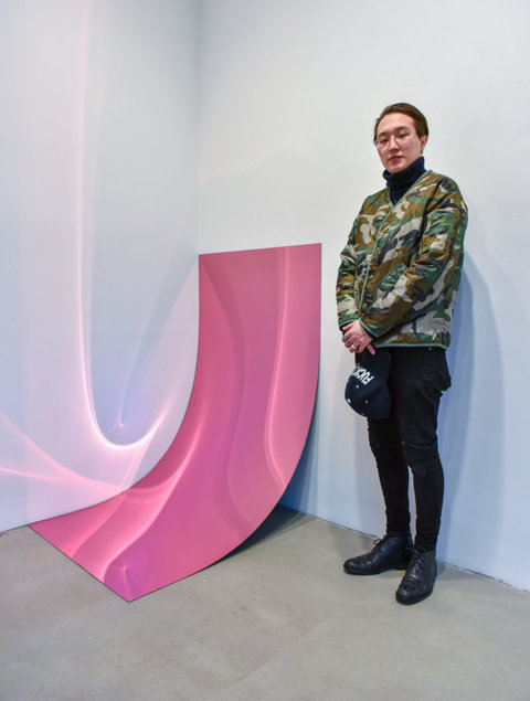 Installation image of an object in the exhibition. The object, on the left, features a pink, shiny, almost metal-like rectangle that leans up against the wall. The rectangle is not strong, and therefore does not stand up on its own, so the shape falls into the crack of the wall and the floor creating an organic leaning object. The light hits the shiny pink material and reflects pink slivers of light onto the white walls.  A person, presumably the artist, stands next to the work on the right with their hands crossed over in front of their body. The figure looks at the camera and wears dark pants, a green camouflage jacket, a black turtleneck, and wire rimmed glasses. 