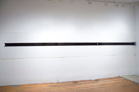 Installation view of an artwork in the exhibition featuring a piece of white paper spanning the length of the wall. Horizontally along the center of the white paper is a thick black line.  