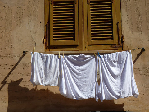 Image of three white sheets hanging on a clothes line in front of a tan building. The shadow of the sheets hits the building just below and above the sheets are two swinging windows with slits up and down them for air and sunlight. The window is light brown and made of wood. 