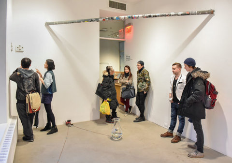 Installation image of the exhibition featuring groups of visitors standing around the room looking at the art and engaging with one another. 