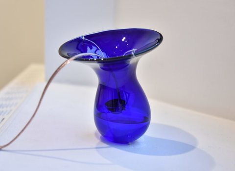 Close-up image of a blue vase-like shape with a rounded bottom and open head. The blue is deep, yet transparent because it is made of glass. Coming out from the top of the blue vase, is a long string like element that travels down and off to the left side of the image. 