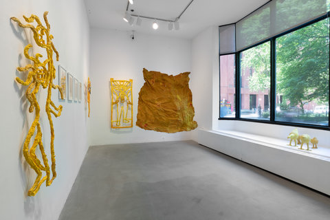 Installation image of the exhibition featuring a large window on the right that allows light to pour in. In the middle of the room, hanging from the ceiling is a large, what seems to be paper mache, or cardboard, crinkled sheet in an orange-yellow color. It hangs off the ground above two feet. From this camera angle, there are multiple works on the left wall, and back wall. On the ledge in front of the right-hand window, sit yellow paper mache birds. On the left organic, abstract paper mache shapes hang on the wall. In the background, mounted to the wall is a yellow paper mache object.  