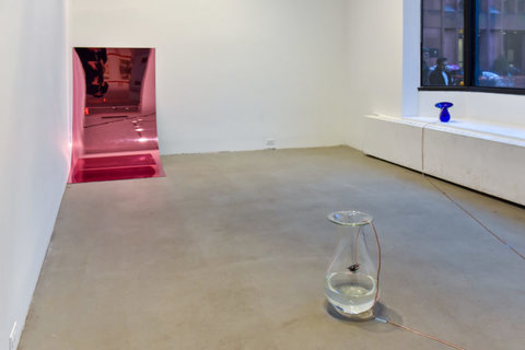 Installation image of several works in the exhibition. On the back wall, a shiny pink, metal sheet leans against the white wall. 