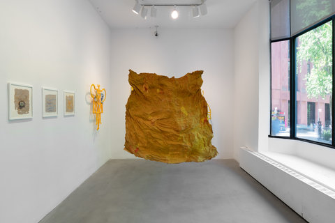 Installation image of the exhibition featuring a large window on the right that allows light to pour in. In the middle of the room, hanging from the ceiling is a large, what seems to be paper mache, or cardboard, crinkled sheet in an orange-yellow color. It hangs off the ground above two feet. On the left side of this hanging object, five pieces of art are mounted on the wall, including four, white framed works and another yellow paper mache work- the works are illegible from this camera angle. 