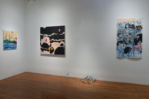 The image shows the corner of a gallery room. On the left wall there is a colorful small to medium-sized landscape painting with many different vibrant colors. On the right wall there are two paintings placed around a sculpture that is centered on the floor against the wall. The painting to the right is a large square canvas painted abstractedly with mostly dark purple and peach. The painting to the right is rectangular but of a similar size. This painting features the repeated profile of a man and the full form of a woman in a sketched style with a blue graffiti style background. In the middle of the right wall there is a pile of corks on the floor. On top of these corks are two flat blue ceramic forms.