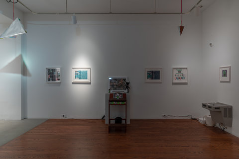 Installation image of the exhibition, from left to right, featuring a metallic pyramid mobile hanging from the ceiling, casting a harsh shadow on the wall behind it. On the back wall, two images are framed with white frames and hung on the wall. The first image is not lit with a spotlight, but the second image that is mostly blue, is lit with a spotlight. In the middle of the wall, a table and pedestal hold several bottles and boxes of Robitussin cough syrup, a ouija board, and two photographic images. After the table, on the right two images are mounted on the wall in white frames. On the right side wall, a white framed image hangs above a silver water fountain. 