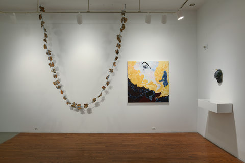 This gallery room has a wooden floor and white walls. Hanging in the middle of the far wall from the ceiling is a chain of ceramic forms. To the right of this U-shaped hanging chain is a square painting featuring mostly yellow, white, and blue. On the perpendicular white wall there is a display case we only see the outside of. Above the display case is a ceramic steel-colored hat hanging on the wall.