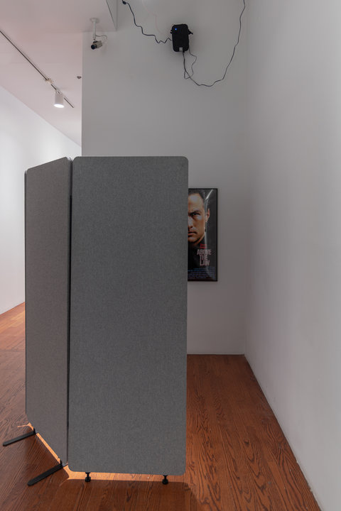 Image of a section of the installation featuring the outside view before entering a cove made from a grey room divider. Peaking out from inside the cove is half a poster featuring a half of a mans face and the words "ABOVE THE LAW" from a movie poster. 