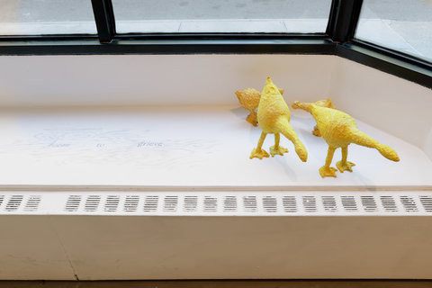 Close-up image of the installation featuring paper mache birds, in yellow, placed on a window bench in the exhibition room. On their left, a sketch of writing states, "ROOM TO GRIEVE."