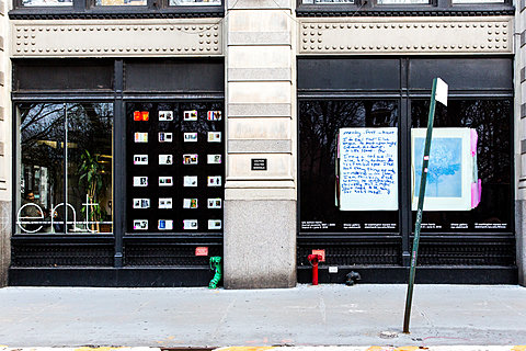 A street-level window display. Two windows are visible: one has a grid of 24 notebook pages against a black background. The other has a zoomed in detail of one notebook page.
