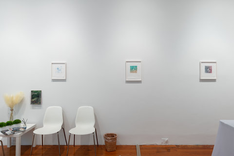 Installation of a section of the exhibition featuring a model waiting room. The waiting room area is complete with two white chairs, a white table with plants and magazines, and four framed works of art on the white wall. 