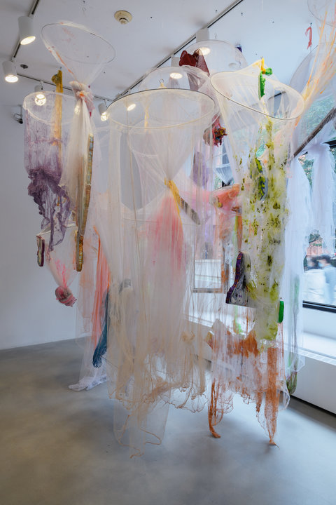 In this floor to ceiling image of a gallery room we see a large hanging installation. The installation is made up of  mosquito nets, tule, and foam cutouts. The mosquito nets are torn in some places and tied together in others. The bottoms of these nets are painted with colors such as purple, blue, green, and orange. There are some found objects tangles into the nets and the nets are of varying lengths. 