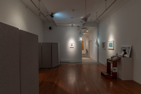 Installation image of a dark room featuring light only from three spotlights. The room includes several smaller installations and also includes grey dividers to separate the sections. 