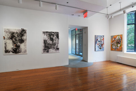 This image shows the corner of a gallery room. On the left wall there are large abstract black and white paintings. Beside them to the right is an open doorway that marks the entrance to this gallery room. On the other side  of the door frame is a rectangular painting that is mostly blue and is close to the corner where it meets with the other wall. On the right wall there is another painting close to the corner that is more square and features the color orange. 