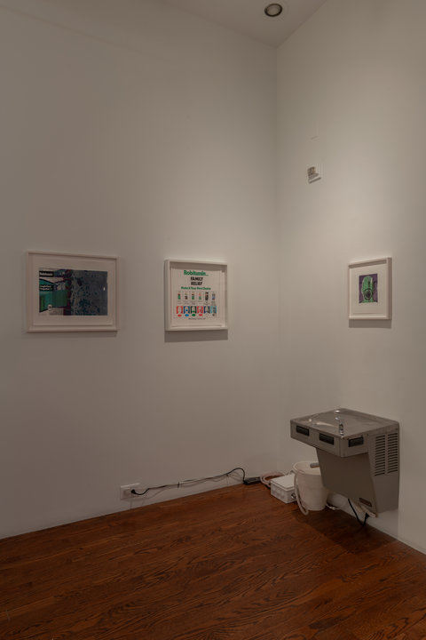 Image of the section of the installation featuring two walls that converge in the corner. On the left wall, two pictures hang in two white picture frames. On the right, one picture hangs in a white picture frame mounted above a water fountain. 